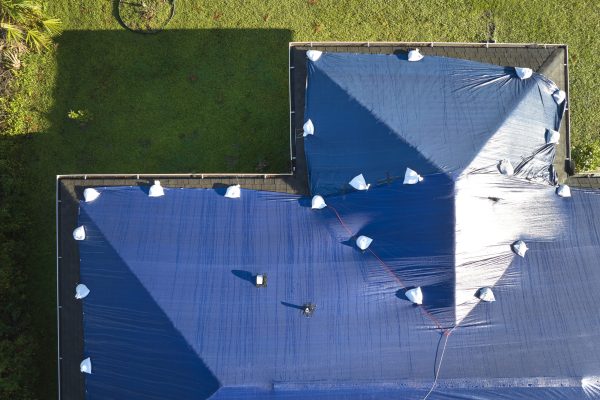 Aerial view of damaged in hurricane Ian house roof covered with blue protective tarp against rain water leaking until replacement of asphalt shingles.